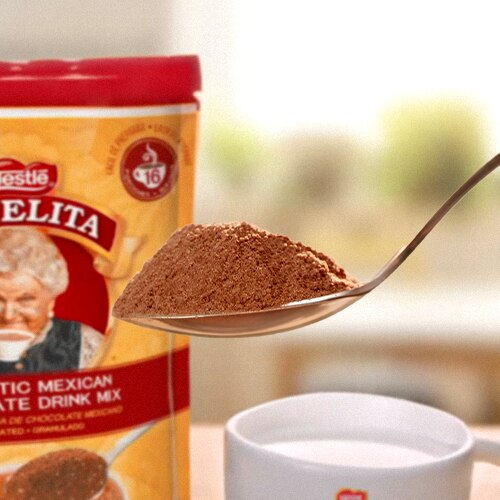 A spoon scoops hot chocolate powder from ABUELITA granulated packaging to a mug of hot milk.