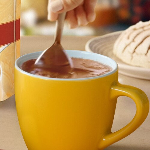 A spoon stirs hot chocolate in a mug in front of granulated packaging and a concha.