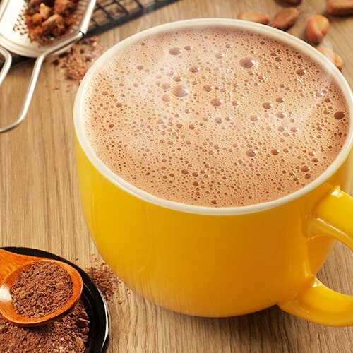 A yellow mug of frothy hot chocolate beside spoons filled with chocolate powder.