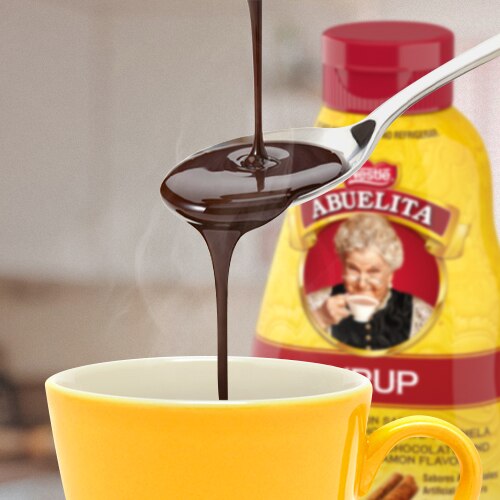 Chocolate syrup pours onto a spoon and into a mug beside a bottle of ABUELITA syrup.
