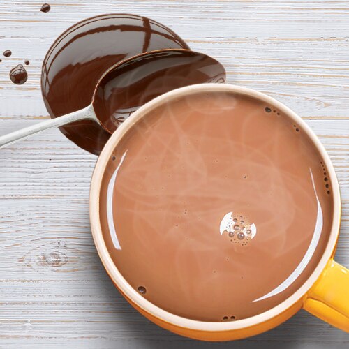 Una taza color amarillo de chocolate caliente junto a una cuchara colocada sobre un poco de syrup de chocolate sobre una mesa de madera. Una taza color amarillo de chocolate caliente junto a una cuchara colocada sobre un poco de syrup de chocolate sobre una mesa de madera.