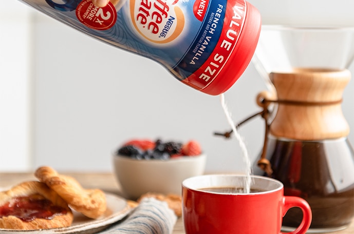 Powder creamer swirls into a red mug beside a bowl of fruit, a pour over coffee maker and a plate of toast.