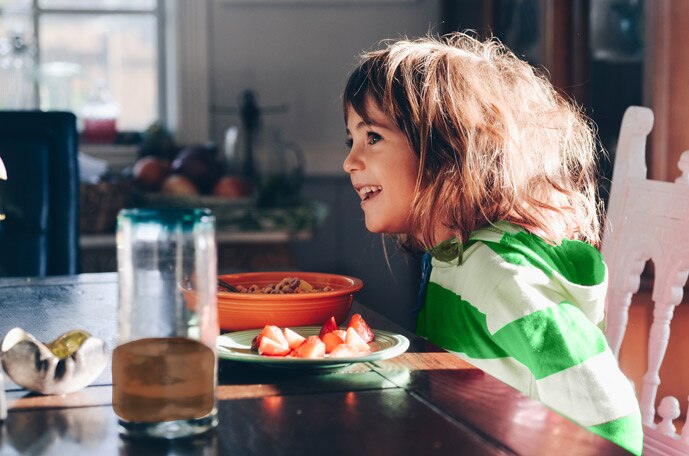 A young kid sits at a kitchen table with a bowl of fruit and a glass of MILO.