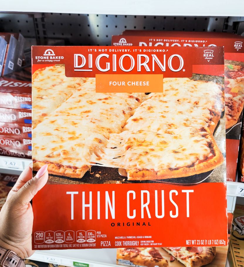 A hand holds a package of DiGiorno Four Cheese Thin Crust pizza in front of a freezer.