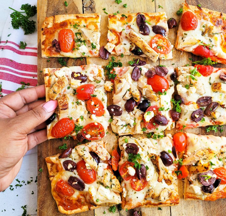 A hand reaches for a DiGiorno Thin Crust pizza topped with chicken, feta cheese, kalamata olives and cherry tomatoes.
