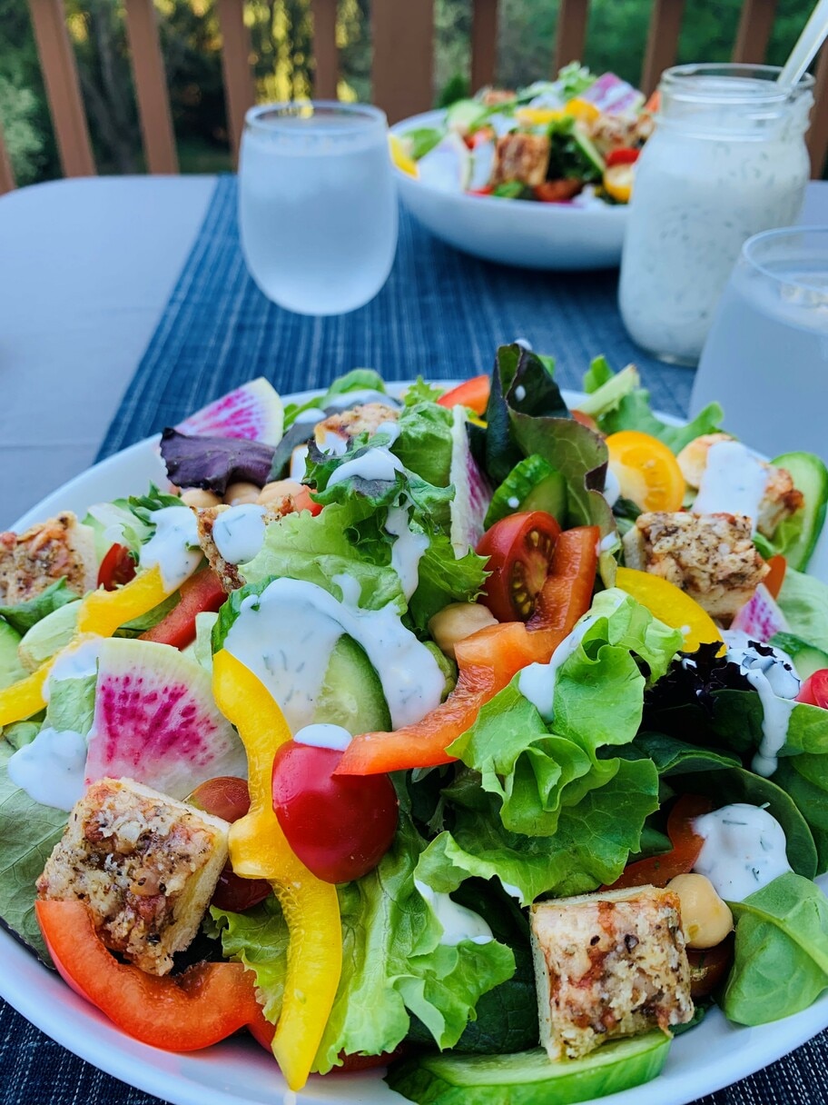 A patio table set with a jar of dressing, glasses of water and plates of salad with peppers, tomatoes, cucumbers and greens.