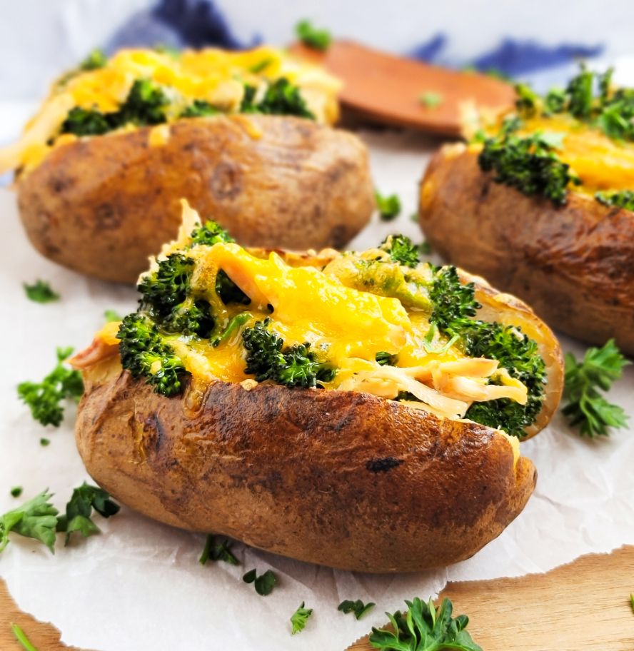 Tres papas al horno repletas de queso, pollo y brócoli sobre una tabla para cortar con papel pergamino.