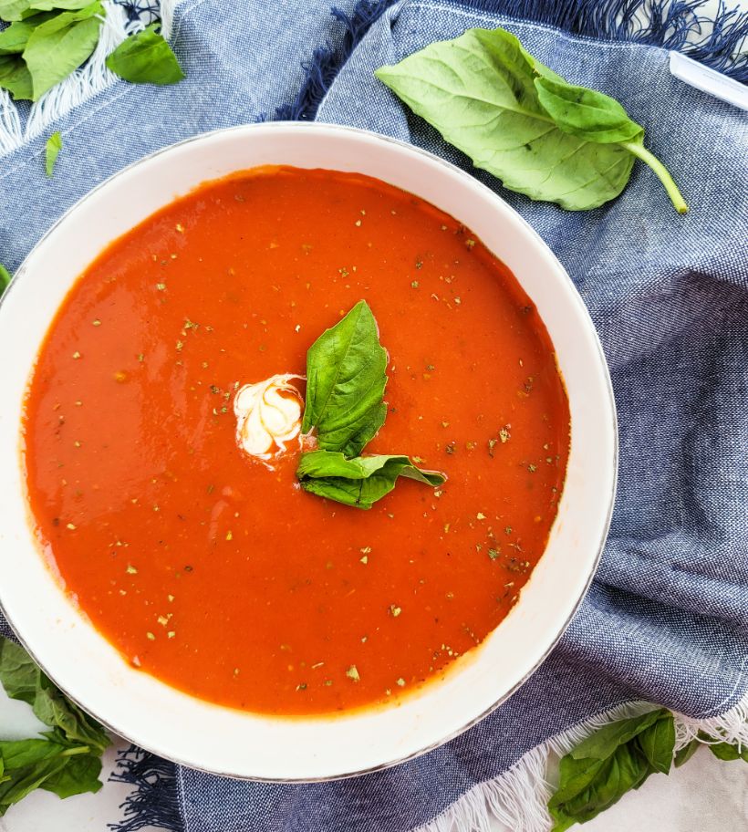 Un tazón de cremoso bisque de tomate decorado con albahaca rodeado de hojas de albahaca y una servilleta de tela.