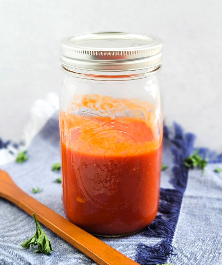 Un frasco de salsa de tomate sobre una servilleta de tela cerca de una cuchara de madera y hierbas de olor esparcidas.