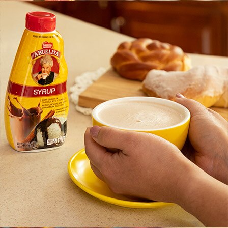 Hands grasp a mug of Mexican hot chocolate near a bottle of Abuelita® syrup and sweet bread.