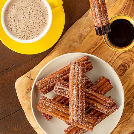 A bowl of churros on a cutting board beside a mug of Abuelita® and a cup of dipping chocolate.