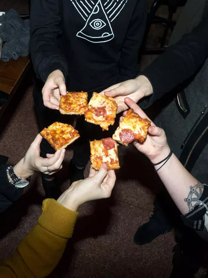 Foto tomada desde arriba de cinco amigos que brindan con rebanadas cuadradas de pizza con cuencos de pepperoni.