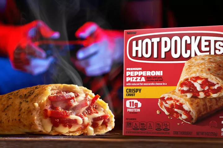 A young man plays video games on the couch behind a box of Pepperoni Pizza Hot Pockets and a Hot Pockets cut in half.