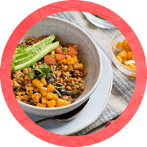 A bowl filled with grains, avocado, tomato, and mango on a white plate and cloth napkin beside a clear bowl of mango surrounded by a red textured circle. A bowl filled with grains, avocado, tomato, and mango on a white plate and cloth napkin beside a clear bowl of mango surrounded by a red textured circle.