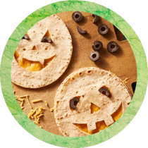 Circle cropped image of Halloween quesadillas with jack-o-lantern faces cut into tortillas and black olives and shredded cheese scattered around surrounded by a green textured circle. Circle cropped image of Halloween quesadillas with jack-o-lantern faces cut into tortillas and black olives and shredded cheese scattered around surrounded by a green textured circle.
