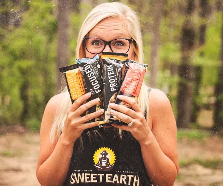 A woman in a black apron with the Sweet Earth logo fans out four frozen burritos in front of her face.