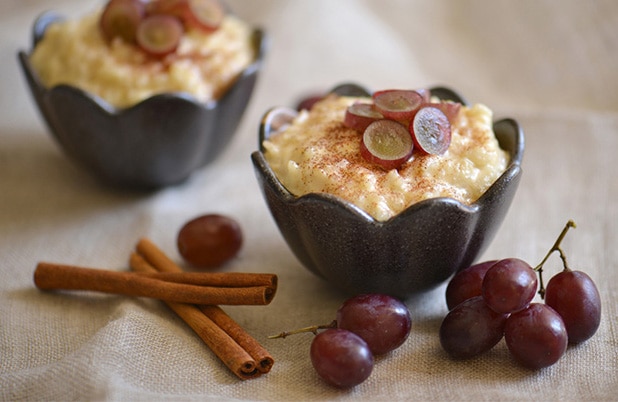 Black bowls filled with Chilean rice pudding topped with sliced grapes besides cinnamon sticks and whole grapes.