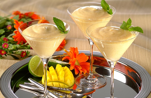 Martini glasses filled with mango mousse on a silver tray with a mango and spoons.