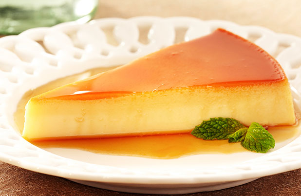 A triangular slice of flan on a white plate with a pool of caramel and mint garnish.