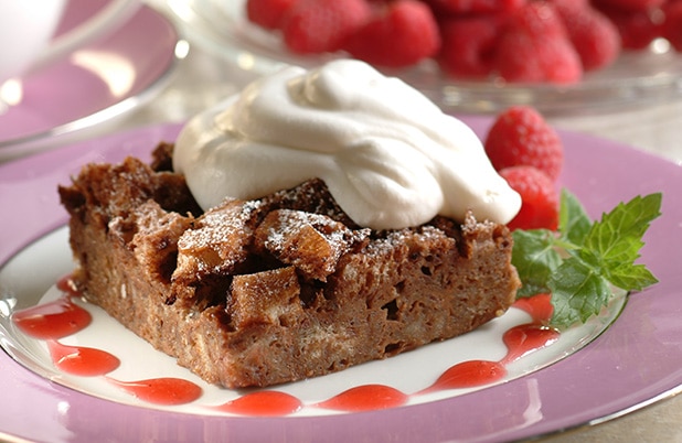 A square slice of mocha bread pudding on a plate with whipped topping and fresh raspberries.