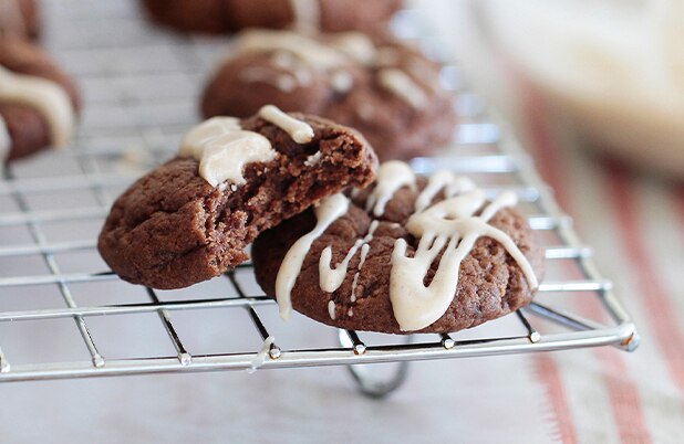 Two chocolate cookies drizzled with white icing on a cooling rack.