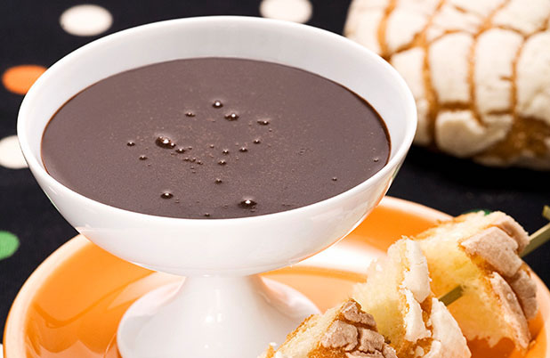 A white dish of chocolate fondue beside skewers of sweet bread.