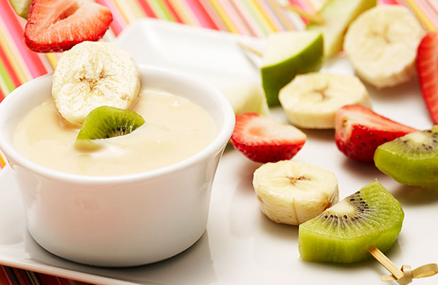 Skewers with banana, kiwi, strawberry and apple on a plate beside a cup of dipping sauce.