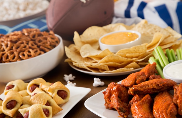 A Game Day spread with bowls of pretzels, chips and queso, pigs in a blanket and wings beside a football.