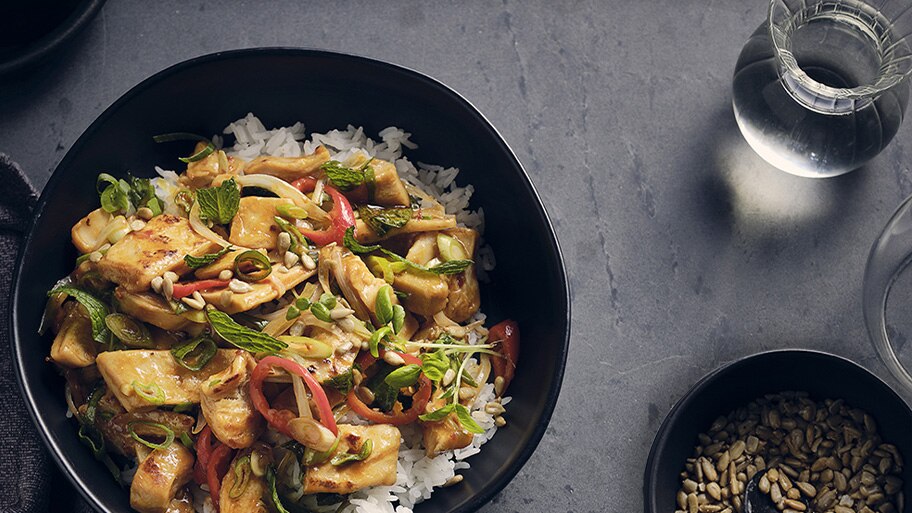 A bowl of Mindful Chik'n stir-fry garnished with green onions and sunflower seeds near a glass of water and bowl of seeds.