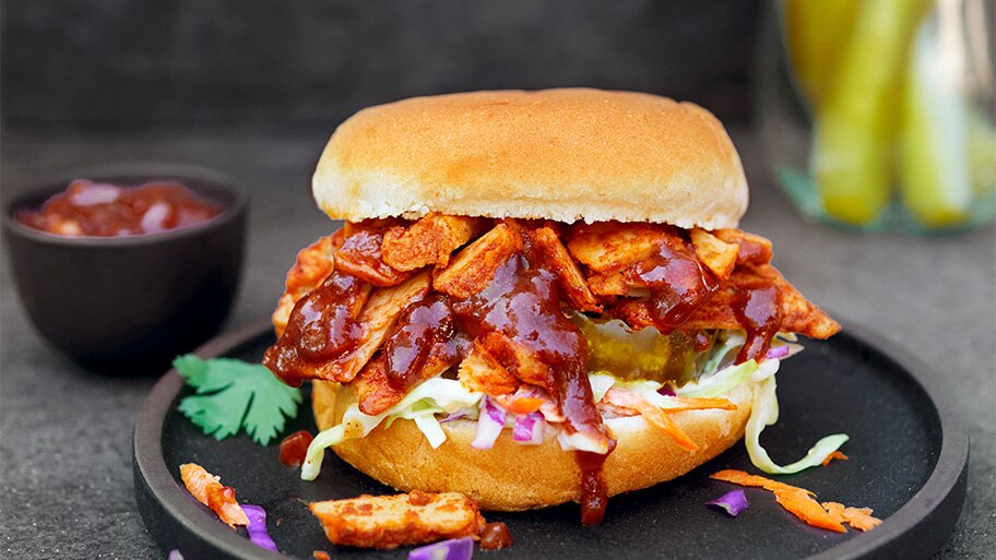 A jar of pickles and cup of ketchup behind a pulled chik'n slider with coleslaw on a black plate.