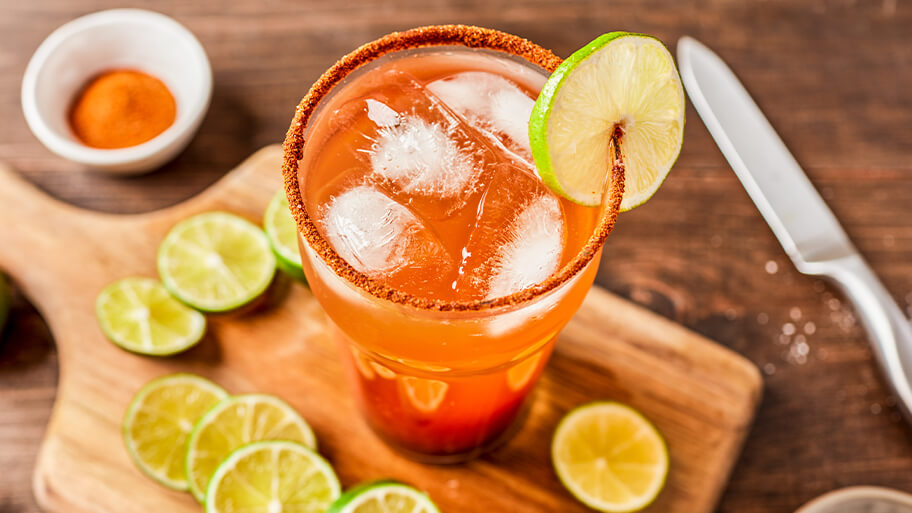 Imagen desde arriba de un vaso de michelada con el borde empolvado con chile y rodajas de lima sobre una tabla de madera.