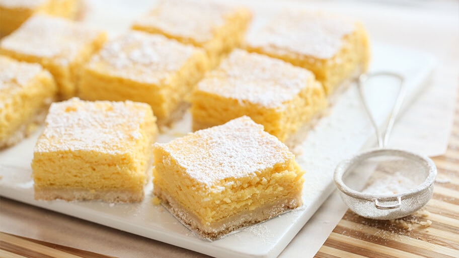 Creamy lemon bar with fresh powdered sugar on top on a marble cutting board with flour sifter.