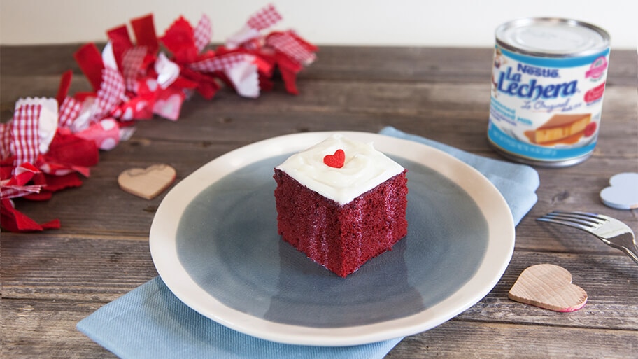 Red velvet poke cake with white cream cheese frosting with a red candy heart on a table with a can of La Lechera sweetened condensed milk.