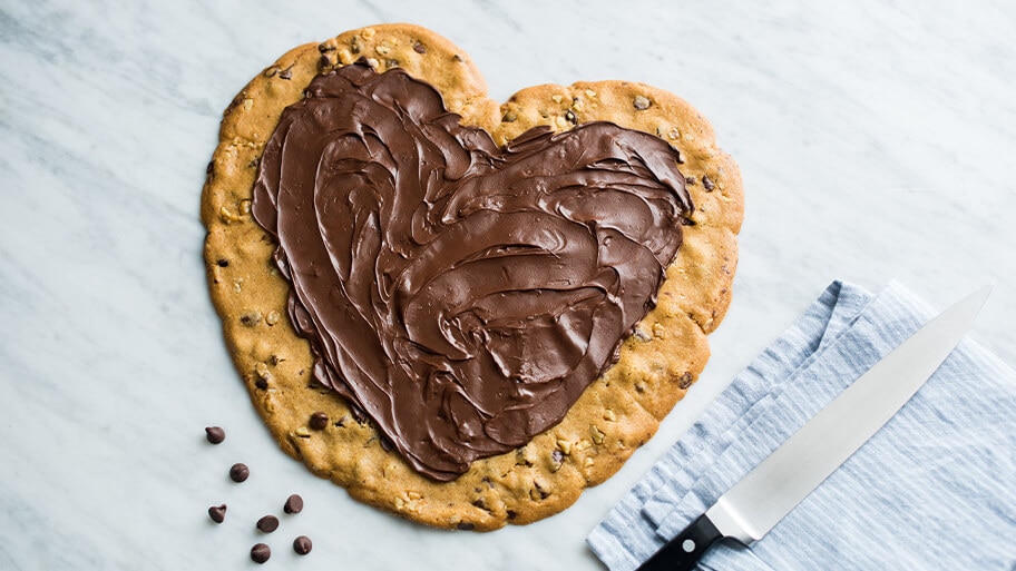 A giant heart-shaped chocolate chip cookie with chocolate frosting in the center on a marble table with knife for cutting.