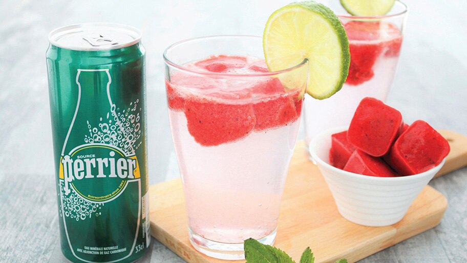 A can of Perrier by a bowl of watermelon ice cubes and glasses of Perrier spritzers with lime garnish on a table.