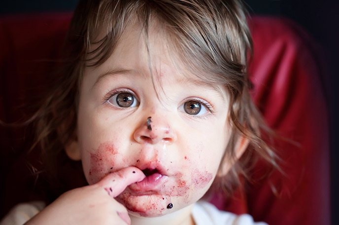 A young girl with her fingers in her mouth and berries smeared on her face.