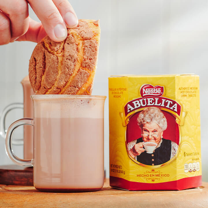 Una mano sumerge una concha dentro de un vaso de leche con chocolate y coco junto a un empaque de tabletas ABUELITA.