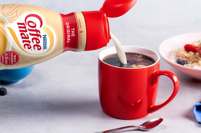 Una botella vierte creamer en una taza roja de café junto a un tazón de granola.