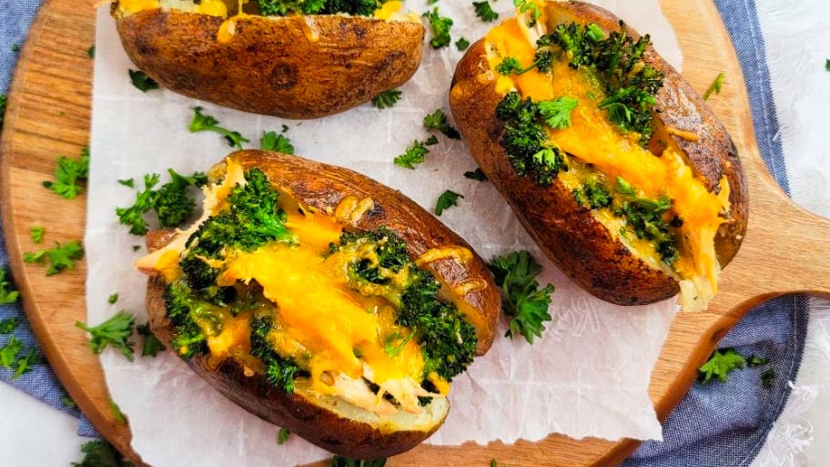 Three baked potatoes loaded with cheese, chicken and broccoli on a round cutting board with parchment.