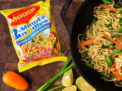 A package of instant noodles near green onions, lime and peppers near a skillet filled with noodles, carrot and peas.