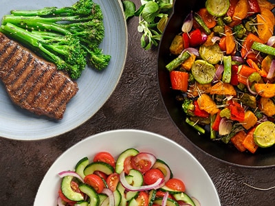 Platos de bistec asado y brócoli, ensalada de tomate con pepino, y un sartén con zanahorias, calabaza y verduras cocidas.