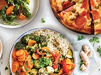 Tazón de verduras frescas y cereales junto a una pizza de pepperoni y una ensalada verde con tomates sobre un mantel blanco.