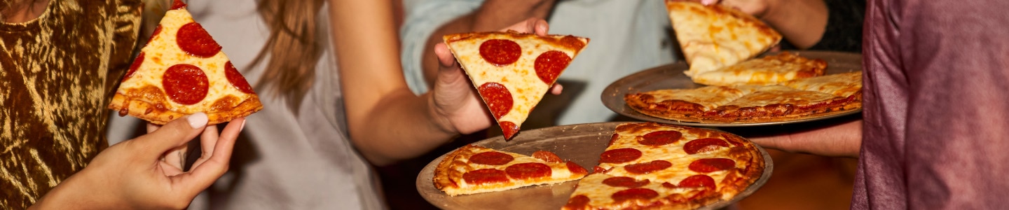 Pepperoni and cheese pizza on platters with people holding slices in the background.
