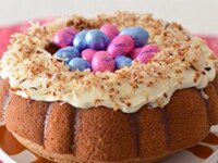 Bundt pan shaped carrot cake topped with icing and toasted coconut, center is filled with pink & blue Easter egg candies.