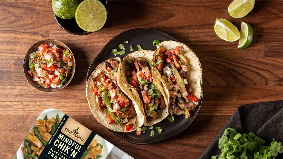 A plate of Chik’n tacos near a bowl of salsa, limes, cilantro and a package of Sweet Earth Mindful Chik’n.