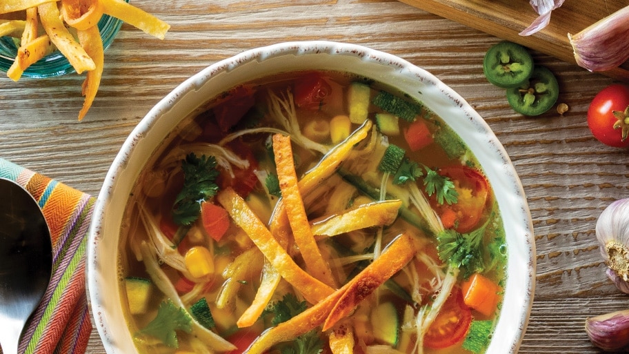 Bowl of chicken vegetable soup with tortilla strip garnish near a spoon, garlic, tomatoes, onions and jalapeños on a table.