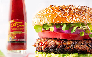 Photograph: sesame-seed burger with lettuce, tomato, onion and microgreens; chili sauce bottle nearby.