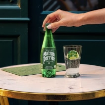 Photograph: hand removing cap from small green Perrier bottle beside branded glass on marble table.