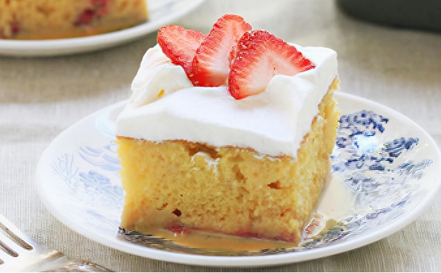 Photograph of a square sponge cake topped with whipped cream and sliced strawberries on a blue plate