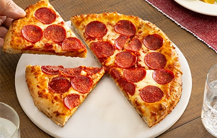 Una mano que agarra una porción de pizza de pepperoni de un plato blanco con una pizza entera.